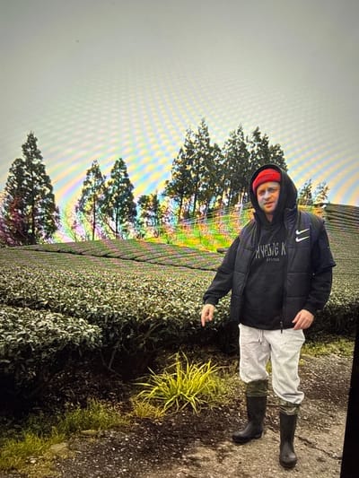 The image depicts a man standing in what appears to be a tea plantation. He is wearing a red beanie, a black hooded jacket over a black hoodie with white lettering, grey sweatpants, and black rubber boots. The man is pointing towards the left of the frame with his right hand. The background shows rows of green tea plants extending up a hillside, with several evergreen trees lining the top of the plantation. The sky is overcast and has a rainbow-like chromatic aberration effect across it. The lower portion of the image shows the ground covered with dirt and some patches of grass. Without additional information, it is impossible to determine if this location is in Taipei, Taiwan, or any other city.