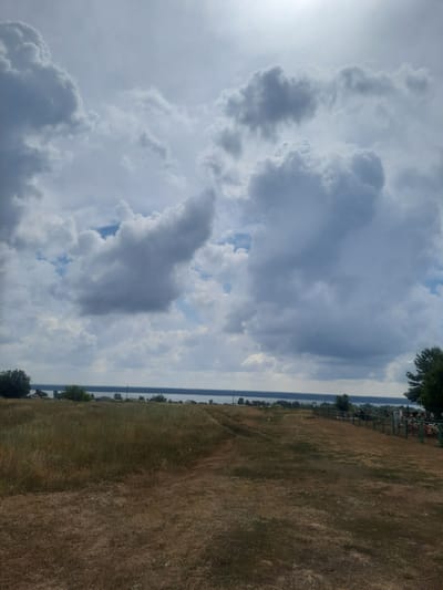 The image depicts a wide landscape under a cloudy sky. In the foreground, there is a dry, grassy field with a dirt path winding through it. The middle ground features a body of water, likely a river or lake, with a distant treeline and some scattered buildings along the shore. The sky is dominated by large, dramatic clouds, with patches of blue sky visible. There is a fence running along the right side of the middle ground, with some structures or objects behind it. No people or animals are visible in the image. The overall impression is one of a rural or semi-rural setting, possibly a cemetery, given the fence and potential markers visible behind it. The location is identified as Крестьянка, Russia.