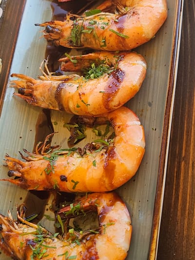 Four large, grilled prawns are arranged vertically on a rectangular, muted green ceramic plate. The prawns are cooked to a vibrant orange hue with visible grill marks and a glossy, dark sauce drizzled over them. They are garnished with finely chopped green herbs, likely parsley or chives. The plate rests on a dark wooden surface, suggesting a table setting. The lighting is bright, creating highlights on the prawns and their sauce. There are no people visible in the image. The scene appears to be a close-up food photograph taken in Jošice, Montenegro.