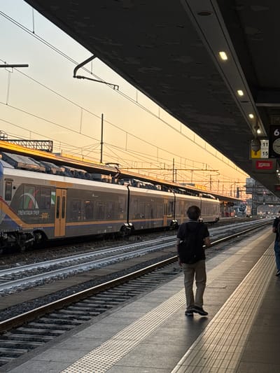 The image depicts a train station platform in Bologna, Italy, during sunset. A Trenitalia Tper train, colored gray and orange with a green and red logo, is stopped on the tracks. A man with a black backpack and khaki pants stands on the platform, facing away from the camera, looking towards the train. The sky is a warm orange hue, indicative of sunset. Overhead, a dark canopy with lights shelters the platform, and electrical wires for the train are visible. Other trains and parts of the station infrastructure are discernible in the background. The platform has textured tiles and a warning strip along the edge.
