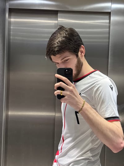 A selfie features a man wearing a white T-shirt with red and black trim and a gold bracelet and ring. He holds a black smartphone in his right hand. The background appears to be a metal elevator interior, visible behind his head and torso. The image is rotated counterclockwise. The man has dark hair, light skin, and a beard.