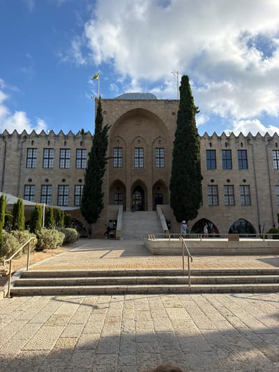 This image captures a view of a stone building with a distinctive crenellated roofline, characteristic of historical architecture. The building appears to be several stories high and features numerous windows, some arched. Two tall, slender cypress trees stand prominently in front of the structure, flanking an entranceway. A flag, yellow and green with some white elements, flies from a flagpole to the left of the building. In the foreground, a wide stone staircase with railings leads up towards the building. Lush green bushes and smaller plants line the sides of the staircase. The sky above is a bright blue, dotted with fluffy white clouds, suggesting fair weather. The sunlight casts shadows on the stone steps, indicating it is likely daytime. The orientation of the image is portrait, with the scene appearing to be viewed from a slightly upward angle. While some people are visible near the entrance and on the staircase, their faces are not clearly discernible. There is some text visible on a plaque near the entrance. The scene conveys a sense of historical significance and serene daytime activity.