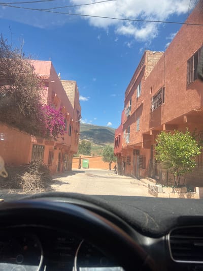 The image, taken from inside a vehicle, shows a street scene in Ouaouizert, Morocco. The street is flanked by peach-colored buildings with barred windows and brickwork. A green tree and a flowering purple bush are visible on either side of the road. A mountain range appears in the distance. The sky is blue with scattered clouds. Part of the car's dashboard and steering wheel are visible in the foreground. Two people are walking in the distance on the right side of the street.