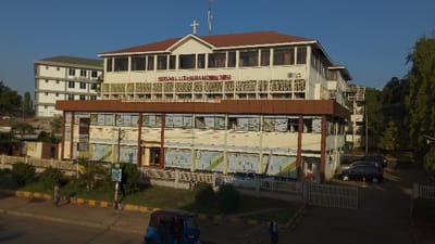The image showcases the Moshi Lutheran Center, a multi-story building with a distinct architectural design. It features a light-colored facade with brown accents, particularly on the roof and window frames. The building displays the text "MOSHI LUTHERAN CENTER" across its front. A secondary, white building is visible in the background. The foreground includes street elements, such as an auto rickshaw, pedestrians, landscaping, and parked cars near a gated entrance.