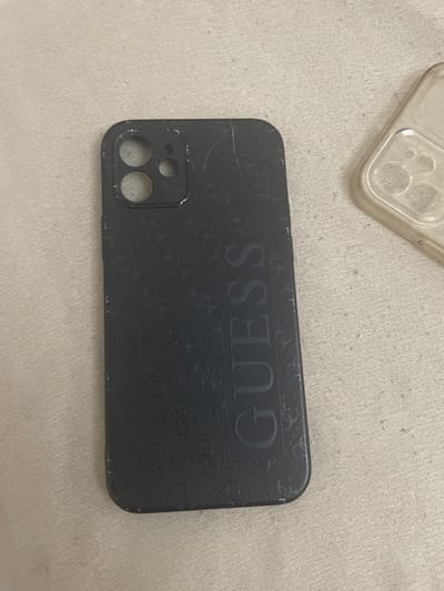 A dark gray smartphone case, featuring a vertically oriented embossed "GUESS" logo and a repeating monogram pattern, rests on a light beige, finely textured fabric surface. The case is designed with cutouts consistent with an iPhone's dual-camera system, including two larger circular openings and one smaller one, and exhibits visible surface scratches, scuffs, and minor abrasions along its edges and surface. Partially visible in the upper right of the frame is a second, transparent smartphone case, also displaying camera cutouts, positioned on the same fabric background.