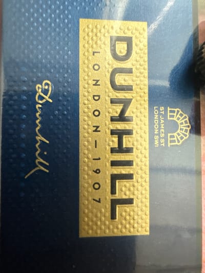 The image shows a close-up view of a dark blue textured surface with gold lettering. The lettering runs vertically and reads "DUNHILL LONDON 1907". Below this, also in gold, are the words "ST JAMES ST LONDON SW1" and a logo that appears to be an archway. To the left of the main gold lettering, there is a script-style gold signature that reads "Dunhill". The surface has a patterned texture of small raised dots. The image does not contain any elements that would allow for the identification of a specific location in Shumentsi, Bulgaria.