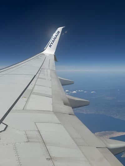 The image is an aerial view from an airplane window, looking out over the wing. The wing is white and appears to be made of metal panels. The Ryanair logo is visible on the wingtip. Below the wing, the ocean stretches to the horizon, with some land visible in the distance. There are a few small clouds scattered across the sky. The image does not contain any information about the city of Ronse, Belgium, or any people.