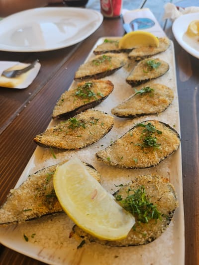 A close-up, eye-level shot shows a rectangular white platter of baked mussels. The platter is filled with what appears to be coarse salt, and the mussels are arranged on top of it. Each mussel is topped with a golden-brown breadcrumb coating and sprinkled with chopped green herbs. Several wedges of lemon are placed around the mussels, one prominently in the foreground. The platter rests on a dark wooden table. In the blurred background, a white plate, a red can, and some white paper are visible. The scene is set in Jošice, Montenegro.