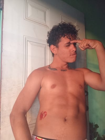 In La Guardia, Venezuela, a shirtless young man with curly dark hair stands in front of a white door. He has a tattoo on his chest that reads "2005" and another on his left side of what appears to be a red animal silhouette. His right arm is flexed, and his left arm is bent at the elbow, with his finger touching his forehead. The background includes a partially visible green wall and some plants. The lighting suggests an indoor environment, likely a home.