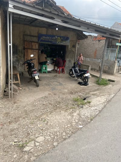 The scene is located in the city of Margaasih, Indonesia at the coordinates -6.965 latitude, 107.542 longitude. There is a small shop on the left side of the image with a sign that reads "Multi Cell Service - Software". Two men are seated inside the shop and a third man stands outside. On the right side of the image, a man stands against a wall with his arms crossed. Two motorbikes are parked in front of the shop. There is a small, wooden, stool in front of the shop. The ground is dirt and gravel. There is a small, paved road on the right side of the image. The sky is overcast. There is a street sign in the distance on the right side of the image. The shop has a metal awning over the entrance. The shop has a metal door and wooden door, both open. The men are wearing casual clothes and are likely customers of the shop. The scene is typical of a small business in a residential area of Indonesia. The scene is peaceful and mundane.
