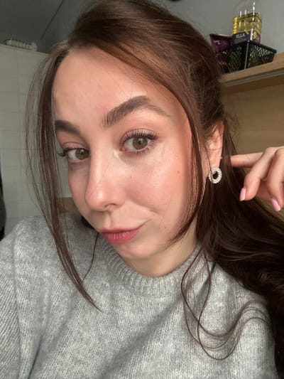 A young woman with light skin, brown hair, and light brown eyes is facing the camera. She is wearing a light gray sweater and a round, sparkling earring. Her fingernails are painted a light pink. It is impossible to determine the location.