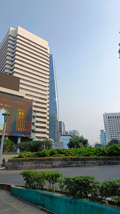 The image captures a view of a modern cityscape with several tall buildings under a clear sky. In the foreground, a paved road curves upwards, bordered by green landscaping and a teal concrete barrier. A pedestrian pathway is visible to the left, where a person on a bicycle is seen.
The dominant structures in the background include a large, light-colored building with a tiered facade on the left, and a sleek, blue glass skyscraper with a distinctive pointed roof in the center. To the right, another white building with arched windows is visible. Several other smaller buildings and modern architecture fill the background.
A large billboard or digital screen displaying a colorful graphic is mounted on the side of the light-colored building on the left. Below the billboard, a sign with "BHRE" is visible, suggesting a commercial area. Lush green plants and trees are strategically placed throughout the scene, creating a sense of urban greenery. The lighting indicates it is likely daytime, possibly morning or late afternoon, given the soft illumination and clear sky. The overall impression is one of a bustling, contemporary urban environment in South Jakarta, Indonesia.