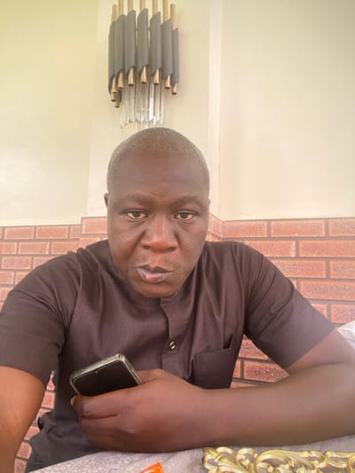This is a portrait of a Black man, who appears to be in his late 40s or 50s. He is wearing a dark brown, short-sleeved button-down shirt. The man is holding a dark-colored smartphone in his right hand, with the screen facing downwards. His gaze is directed towards the camera, and his expression is neutral.

The background features a brick wall with reddish-brown bricks laid in a running bond pattern. To the left of the man, there's a cream-colored wall with a light fixture or decorative element that appears to be made of metal tubes and clear rods. To the right of the man, a decorative gold frame is partially visible. The overall setting suggests an indoor environment, possibly a home or a common area. The lighting appears to be natural, indicating it might be daytime. No specific weather conditions are discernible. The location context of Unguwar Sarki, Nigeria, is provided, but there are no overt visual cues in the image that specifically point to this location. The image has a slightly tilted orientation.