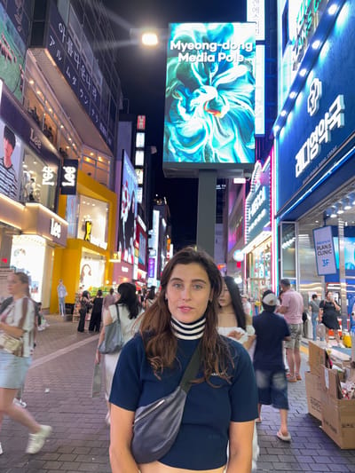 The image shows a street scene at night in Myeongdong, a popular shopping district in Seoul, South Korea. The foreground is dominated by the face of a woman with long, dark, wavy hair and blue eyes, wearing a dark blue long-sleeved shirt with a white and black striped turtleneck. A dark gray crossbody bag is worn over her left shoulder. The background is a busy street with numerous neon signs and advertisements in Korean and English.
To the left, a large digital billboard displays "Myeong-dong Media Pole" with an abstract, colorful image. Below this, a sign for "Lee" fashion store is visible, featuring a model's image. Further down the street, other shopfronts and pedestrian traffic are apparent. People are walking in both directions on a paved walkway. To the right, a blue building with illuminated signage reads "PLAN.S CLINIC 3F" and other Korean text. Several cardboard boxes are stacked on the right side of the walkway, some with visible text and labels, including "OLIVE YOUNG". The overall atmosphere is one of vibrant commercial activity typical of a bustling city center at night.