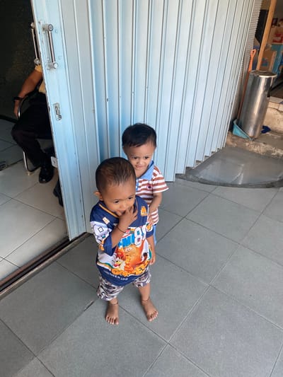 Two young children stand on a tiled patio outside a building with a blue corrugated metal door. The child in the foreground, facing slightly to the right, wears a blue and white shirt with a cartoon character and camouflage shorts. They have their finger in their mouth. The second child, standing behind and to the left, faces forward and wears a brown and white striped shirt. In the background, a person in dark pants and a yellow shirt sits in a chair partially visible through an open doorway. A tall, silver trash can and a broom are also present on the patio. The scene appears to be outdoors in an urban or commercial area, potentially in Ta', Indonesia, given the context.