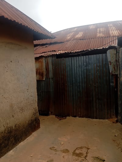 The image depicts an outdoor scene in Dutsen Alhaji, Nigeria. The primary subjects are two adjacent buildings with corrugated iron roofs, showing signs of rust and age. The building on the left has a light-colored, rough-textured wall, stained at the bottom. The building on the right is largely obscured by a wall made of vertically corrugated, rusty metal sheeting. The ground is a mixture of concrete and earth, with some puddles and debris.
The scene appears to be a residential or utility area, possibly a courtyard or alleyway between structures. The overall impression is one of a rural or semi-urban environment, with a sense of wear and tear on the materials.
Notable details include the weathered appearance of the roofing and wall materials, suggesting exposure to the elements over time. The lighting suggests it is daytime, though the sky is overcast, contributing to a muted and somewhat somber atmosphere. There are no people or animals visible in the frame, and no discernible activity is taking place. The location cues are limited to the architectural style and materials common in many parts of Nigeria. There is no visible text in the image.