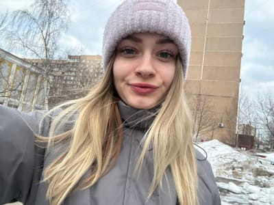 A young adult female is depicted in a close-up, chest-up selfie, positioned outdoors against an urban backdrop on what appears to be a cloudy winter or early spring day. The subject has fair skin, long blonde hair, and green eyes. She is wearing a light lavender knitted beanie with "Really?" inscribed on it and a grey puffer jacket with a high collar. Her lips exhibit light reddish-tinted makeup, and she displays a slight smile. The camera is held at arm's length, suggesting a self-portrait.
The background reveals a street scene with architectural elements and vegetation. To the subject's left, a portion of an older, light-yellow or cream-colored building with visible window frames and stucco details is present. Further behind this structure and centrally located in the mid-ground, a multi-story residential building with a lighter, possibly beige, facade is visible through bare deciduous tree branches. Dominating the right side of the background is a tall, multi-story beige-colored panel apartment building, displaying prominent vertical seams indicative of its construction, with a white air conditioning unit mounted on its facade at a lower level. In the immediate foreground on the right, a substantial pile of dirty, melting snow and ice is visible, consistent with cold weather conditions. The sky above is overcast.
This scene is located in the city of Reutov, Russia. The older, light-yellow building on the left is identifiable as the Children's Art School (Detskaya Shkola Iskusstv) at Pervomayskaya Street, 13. The prominent beige panel building on the right is the residential building located at Pervomayskaya Street, 48. The photograph appears to be taken from an outdoor public area, such as a sidewalk or open space, looking approximately southeast towards these specific landmarks.