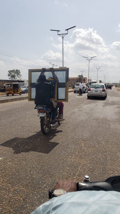 In Hayin Banki, Nigeria, a man is riding a motorcycle on a paved road, precariously carrying a large, rectangular window frame. The frame is held aloft by the man and the motorcycle rider, posing a safety hazard. Other vehicles, including a white car and a yellow auto-rickshaw, are visible on the road. Streetlights are seen along the roadside, and the sky is partly cloudy.