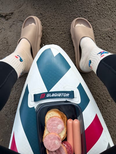 A first-person, overhead view captures a person's lower legs and feet, clad in black leggings, cream-colored socks adorned with colorful embroidered bird-like motifs in blue and red, and light brown chunky slide sandals. The person is positioned on a dark brown, textured ground, consistent with damp earth or muddy sand. Resting on the ground between their legs is the front portion of an inflatable stand-up paddleboard (SUP), primarily white with dark teal/blue and red geometric patterns, including stripes and triangles. A black fabric handle with the brand name "GLADIATOR" and a red "G" logo stitched on it is visible near the board's nose. Placed centrally on the board, in front of the person, is an open rectangular black plastic food container. Inside the container are several slices of white bread, multiple round slices of cooked sausage or salami, and two pink frankfurter-style sausages. The scene depicts a casual outdoor meal or snack break. The image contains no visual information to determine its specific geographical location.