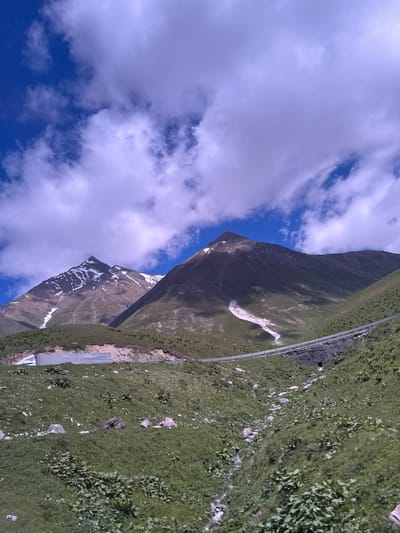 The image shows a mountainous landscape with lush green slopes and snow-capped peaks under a bright blue sky with scattered clouds. A winding road with a metal guardrail is visible cutting across the middle ground, and a large truck trailer is parked on the side of the road. The scene appears to be located in the vicinity of Gudauri, Georgia, a popular ski resort known for its scenic mountain views and challenging terrain.