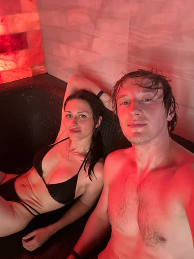 The image shows a selfie of a man and a woman in a spa-like environment. They are both wearing swimwear. The woman is wearing a black bikini and is lying down, with her arm behind her head. The man is also in the frame, looking at the camera. The background features pinkish-red illuminated salt blocks and tiled walls, typical of a salt room or sauna. The lighting is predominantly red, creating a warm and atmospheric glow over the subjects and their surroundings. The context of the image suggests a relaxation or wellness setting. There is no information provided to associate this image with the city of Murino, Russia.