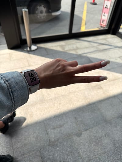 A photograph shows a person's arm and hand, raised to display a peace sign with the index and middle fingers. The fingernails are long and painted with a light color. A smartwatch with a white band is on the wrist, displaying "16:20." The person is wearing a light blue denim jacket. The background consists of gray tiled flooring, a window with sunlight casting shadows, and a glimpse of a vehicle wheel. The letters "ALE" are visible in reverse on a sign seen through the window. The location is Bayrampaşa, Turkey.