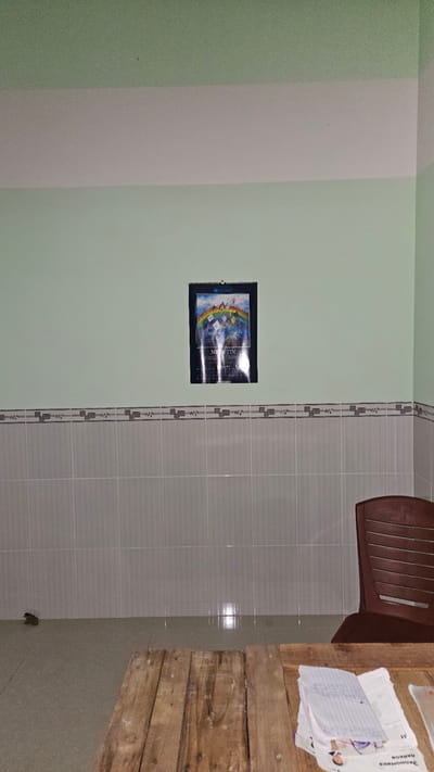 This image, rotated 90 degrees clockwise, captures an indoor domestic space, likely a room within a home.
**Scene/Setting:** The room features contrasting wall treatments. The wall on the left (as viewed in the rotated image) is primarily painted a light, muted green. A vertical, lighter grey or off-white stripe divides this green wall. To the right of this painted section, the wall is covered in light grey rectangular tiles, separated by a decorative patterned border running horizontally across the middle.
**Objects:** A calendar, featuring a vibrant, possibly fantastical illustration (including what appears to be a rainbow or arch), is hung on the light green wall. The word "NEXUS" is clearly visible on the calendar, with "BIORHYTHM" partially seen. In the lower right corner, a rustic wooden table constructed from rough-hewn planks is visible. On this table, several papers are scattered, one containing handwritten notes and another with printed text. The words "Экономика" (Economics) and "данных" (data), written in Cyrillic script, are discernible, along with the letter "M." A brown plastic chair is partially visible, positioned above the table in the upper right. Near the base of the tiled wall, a small, dark, irregular object, possibly a small animal or debris, is present on the floor.
**Activity or Event:** No activity is taking place; the image presents a static view of the room.
**Notable Details:** The lighting in the room appears to be natural or ambient, suggesting daytime. Reflections on the tiled wall indicate a light source. The combination of simple painted walls, tiled sections, and basic wooden furniture gives the space a functional and unpretentious character. The presence of text in Cyrillillic on the papers is a notable detail for a setting in Cam Đức, Vietnam, potentially indicating educational materials or a cultural connection.