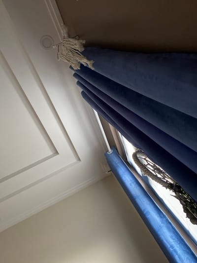 The image shows a close-up of a ceiling and window treatment with blue curtains. The ceiling is white with decorative molding and recessed lighting. The curtains are a deep blue velvet and are partially drawn, revealing a glimpse of a window with what appears to be a decorative rope or netting. The scene is indoors and lit by natural light coming from the window. The perspective is looking upwards from a low angle. There are no people or animals visible in the image. The location of Lâm Viên - Đà Lạt, Vietnam is not discernible from the visual information provided in the image.