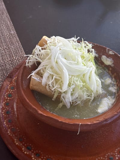 The image shows a serving of enchiladas mineras, a traditional dish from Aguascalientes City, Mexico. The enchiladas are rolled tortillas, possibly corn, filled with cheese or other ingredients, and topped with shredded cabbage, onions, and possibly sour cream or a similar dairy product. They are served in a terracotta bowl filled with a green sauce, possibly a tomatillo-based sauce. The bowl sits on a dark surface, possibly a table, with a textured mat visible in the lower left corner.