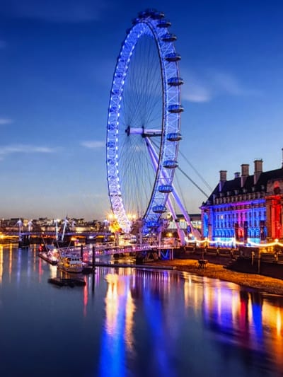 A nighttime photograph captures the London Eye, prominently illuminated in blue, dominating the left side of the frame. To its right, the multi-story County Hall building features several chimneys and is brightly lit with blue, purple, and red artificial lights. These structures line the bank of the River Thames, which occupies the lower half of the image and vividly reflects the blue, purple, red, and yellow light sources from the scene. In the mid-ground, a bridge with illuminated arches spans the river, and several small boats are moored along the near bank. The sky is a deep twilight blue with faint cloud formations. The environment is an urban riverfront at night, located in London, United Kingdom. No people or explicit actions are discernible.