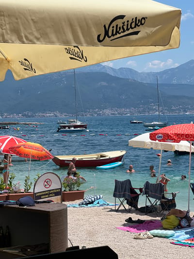 The image depicts a sunny day at a beach in Baošići, Montenegro. In the foreground, a large, yellow umbrella with the "Nikšićko" logo provides shade. The water is clear and turquoise, with several boats visible in the background, including sailboats and smaller motorboats. People are enjoying the water, swimming and relaxing. Beach chairs and towels are scattered on the pebbled shore. A sign with a prohibition symbol indicating no food or drinks is also visible. The backdrop features lush green hills and mountains, characteristic of the Bay of Kotor.