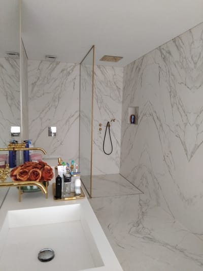 This is a medium shot of a modern, luxurious bathroom. The walls and floor are covered in white marble with grey veining. To the left, a large vanity features a white sink, gold faucets, and a bouquet of orange roses. A mirror reflects the opposite wall. The right side of the image shows a walk-in shower with gold fixtures and a black shower hose. A small recessed shelf holds a blue bottle. The overall impression is one of clean, minimalist design with a touch of opulence. The location is not discernible from the image itself.