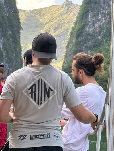 Three people stand on a vessel near Đồng Văn Commune, Vietnam. A person with hair in a bun leans over the railing, wearing a white t-shirt. Another person, facing away from the camera, wears a baseball cap and a tan t-shirt with a diamond logo and the word "Wildrun" on it. A third person is visible between them, and a fourth person is visible in the bottom of the frame. Lush, green mountains rise in the background. The image appears to have been rotated.
