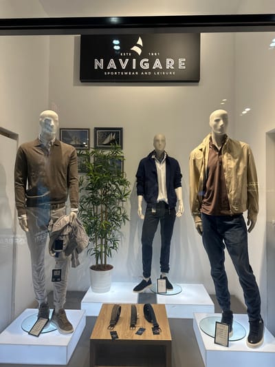 The image depicts a storefront display window of a "NAVIGARE" clothing store. Three mannequins are arranged vertically. The top mannequin wears a tan jacket, brown shirt, and dark jeans. The middle mannequin is dressed in a navy jacket over a white polo shirt with dark jeans. The bottom mannequin wears a brown sweater, light-colored jeans, and has a gray bag draped over its shoulder. A potted plant is situated between the middle and bottom mannequins. The store sign is visible on the left side. The store is located in Novi Sad, Serbia. Belts are displayed on a wooden stand.