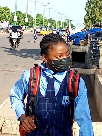 A young Black person wearing a light blue collared shirt, a dark plaid vest, a blue and white tie, and a black mask walks on a paved road in Wuro Dole, Nigeria. They are carrying a red and black backpack. The road is lined with trees and streetlights. In the background, several vehicles are visible, including a motorcycle with a rider, a car, and yellow auto-rickshaws. The scene appears to be a busy street in a Nigerian city.