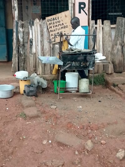The image shows a man cooking in front of a small stand in Kivaywa, Kenya. The stand is made of a wooden fence with a metal frame and a grill. The man is wearing a white shirt and is standing next to the grill.  There is a sign behind him that says "UGALI, MGUU, NYAMA, MATUMBO, KUKU, ML" in a yellow color.  The man appears to be preparing food for customers. The scene is located at latitude 0.562 and longitude 34.805 in the city of Kivaywa, Kenya. There are a number of containers, some of which are full of water. The area is mostly dirt, with a few rocks and some grass.