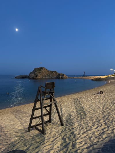 The image depicts a beach scene at dusk in Blanes, Spain. The sky is a deep, clear blue, with a sliver of the moon visible. The foreground is dominated by a sandy beach with distinct ripple patterns, suggesting it may have been recently raked or the tide has receded. A wooden lifeguard chair stands prominently on the sand, casting long shadows.
In the middle ground, the calm blue sea meets the shore, with gentle waves lapping at the sand. A rocky outcrop juts into the water, and several small yellow buoys are scattered in the sea. Further out, the coastline is visible, with buildings and lights indicating a town. A few boats are anchored in the distance.
The beach appears to be populated by a few people. Some are walking along the water's edge, while others are sitting or lying on the sand. One person is visible on the rocky outcrop, and a few figures can be seen in the water. The overall atmosphere is peaceful and serene, typical of a beach at the end of the day. The presence of the lifeguard chair suggests the beach is a recreational area. The lighting indicates it is either early morning or late evening.