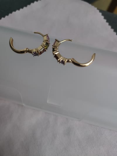 Two gold hoop earrings with inlaid colored gemstones are placed on a white textured surface. The earrings are positioned slightly apart, with the left earring closer to the viewer. The gemstones appear to be purple, green, and possibly pink. The background is a blend of white and a muted blue-gray surface.