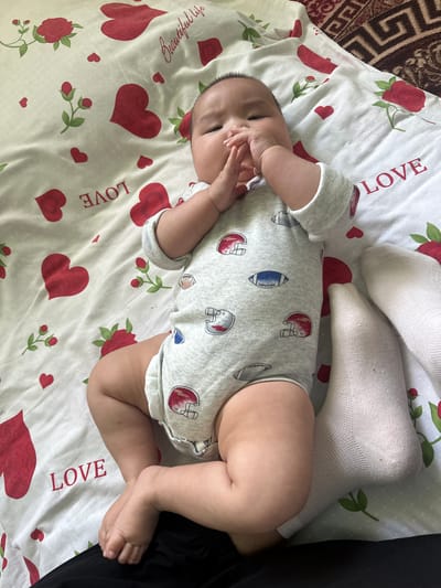 The image shows a baby lying on a white sheet decorated with red hearts and roses. The baby is wearing a grey onesie with a pattern of football helmets and footballs. The baby's hands are clasped in front of their mouth, and their feet are bare. Two white socks are visible to the right of the baby. The background shows a brown and beige patterned rug. The scene is indoors, likely in a bedroom or living room. The location in Тайгараев, Kyrgyzstan is inferred from the general context of similar images, but there are no specific visual cues in this image to confirm this location.