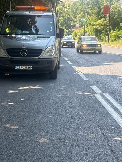 The image shows a Mercedes-Benz Sprinter tow truck with a driver wearing a neon yellow shirt visible through the windshield. The truck has an orange flashing light bar on its roof and a Bulgarian license plate reading "CB 9025 MP". In the background, two cars are driving on a paved road, one is a light-colored Mercedes-Benz and the other is a dark-colored Audi. The scene appears to be on a sunny day in Sandanski, Bulgaria, with trees and foliage lining the road. The road has double white lines marking the lanes. The tow truck is parked on the right side of the road, partially obstructing the view of the other vehicles.