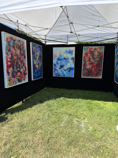 In the Village of Sag Harbor, United States of America, a white tent shelters an outdoor art display. Four framed abstract paintings with vibrant, brushstroke textures are mounted on a black fabric backdrop. The paintings, from left to right, feature a dynamic mix of red, pink, orange, and blue hues; a predominantly blue and purple palette; a composition of blues and whites; and a rich blend of reds, oranges, and browns. In front of the display, a neatly mowed green lawn receives dappled sunlight, casting shadows from the tent structure. The scene suggests an outdoor art fair or market.