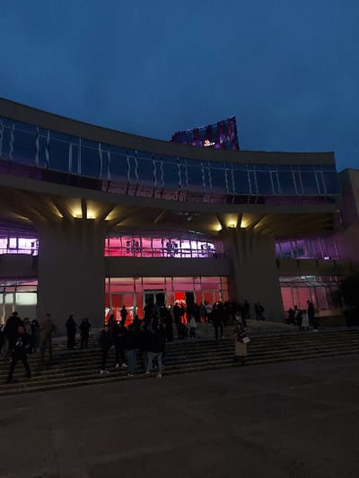 The media file captures a modern, multi-story building illuminated in vibrant pink, red, and purple lights against a deep blue dusk or night sky. The building features extensive glass facades and prominent structural elements. In front, a wide set of steps leads up to the main entrance, where a crowd of people is gathered. Most individuals are seen in silhouette against the bright interior lighting of the building. They appear to be standing or queuing, suggesting a public event or gathering. One person, positioned on the steps, holds a white sign, though the text on it is unreadable.
Visible text includes the partial word "VENU" (likely "VENUE") on a red-lit section of the building's facade. At the very top of the building, another sign glows with blue and purple lights, but its text or logo is indistinct. The artificial lighting creates a dynamic and colorful atmosphere. The scene depicts an active urban setting, likely a cultural or entertainment venue, with people congregating outside during the evening.