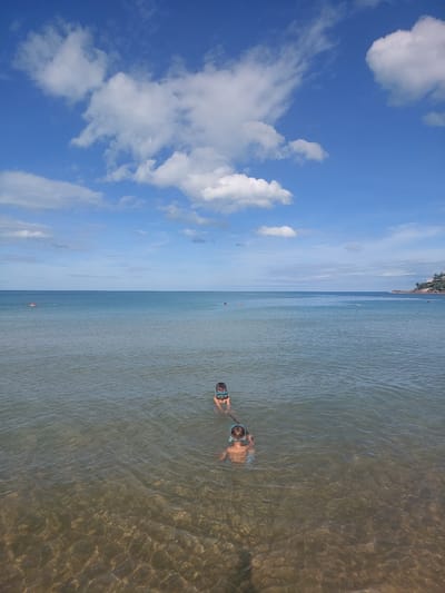 In Baan Chaweng Noi, Thailand, two young children, each wearing a blue diving mask, are swimming in clear, shallow ocean water under a bright blue sky dotted with fluffy white clouds. The water's clarity allows the sandy seabed to be visible. In the distance, other people are scattered across the water, some appearing to be on inflatable rafts or swimming. A faint outline of a coastline with greenery and structures is visible on the right horizon.