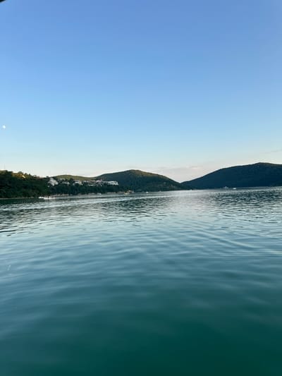 The image depicts a scenic view of a body of water, likely a lake or sea, with a lush, tree-covered hillside bordering it. The water's surface is rippled, reflecting the light and creating a dynamic texture. The hillside is dotted with scattered white buildings, suggesting a settlement or resort area. The sky is a clear, pale blue, transitioning to a lighter hue near the horizon. The overall impression is one of tranquility and natural beauty. A small, crescent moon is visible in the sky. The image appears to have been taken during daylight hours, possibly in the late afternoon or early evening, given the soft lighting. There is no visible text in the image. The location is identified as Abrau-Dyurso, Russia.