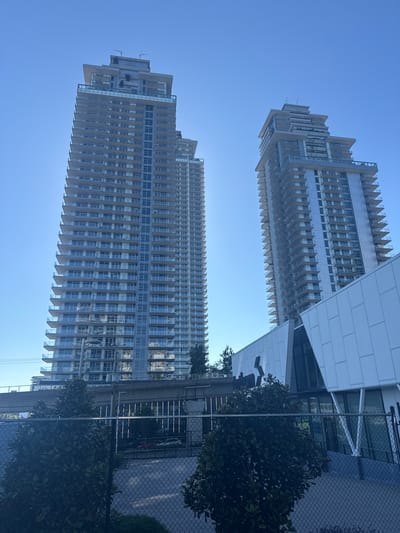 The image captures a scene in Coquitlam, Canada, dominated by two modern, high-rise residential towers against a clear blue sky. The towers feature numerous balconies with glass railings and light-colored facades. A contemporary building with a white, angular exterior and large glass windows is situated in the foreground, partially obscuring the base of the towers. In the middle ground, a concrete structure, likely an elevated transit line, runs horizontally between the towers. The foreground shows a fenced area with manicured trees and bushes. The overall impression is of a well-developed urban environment with a focus on modern architecture.