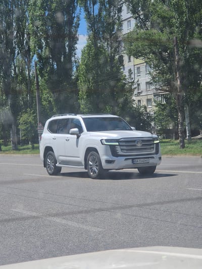 A white Toyota Land Cruiser is driving on a paved road in Kropyvnytskyi, Ukraine. The vehicle is a modern SUV with distinctive LED headlights and a chrome grille. The car has a license plate that reads "BA 5540 HI". In the background, there are tall trees with lush green leaves, a multi-story apartment building, and a utility pole. The sky is blue with scattered clouds. The road has lane markings and some tire marks are visible on the asphalt. A road sign indicating children crossing is visible on the left side of the road. The overall scene appears to be a bright, sunny day.