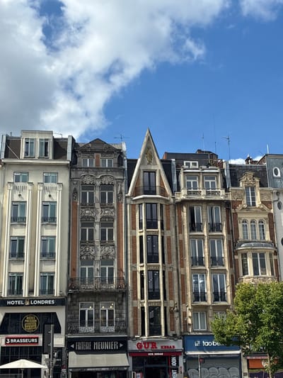 The image captures a street view in Lille, France, featuring a row of multi-story buildings with intricate architectural details. The buildings exhibit a mix of styles, with the leftmost structure being a lighter-colored building with repeating "HOTEL LONDRES" signage and vertical decorative elements. To its right, a darker building displays ornate stone carvings and brickwork around its windows. Further to the right, a building with a pointed gable features a golden bird emblem at its apex. The ground level of these buildings is occupied by various businesses, including "3 BRASSEURS," "FRITERIE MEUNIER," and "GUR KEBAB," indicating a commercial street. The sky above is blue with scattered white clouds. A lush green tree is visible on the right side of the frame. The overall scene depicts an urban environment with a focus on the historical architecture of Lille.