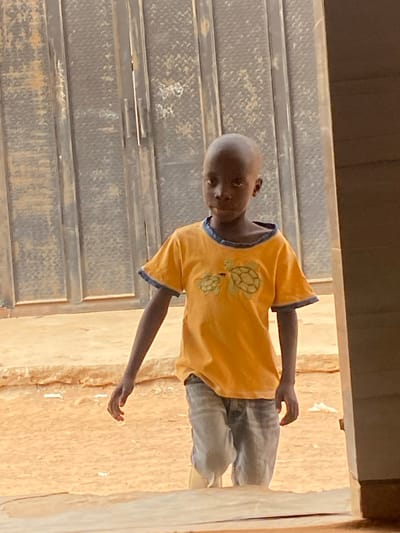A young boy stands, his head bald, gazing downward. He wears a short-sleeved yellow shirt decorated with two sea turtles and blue trim on the sleeves and collar. He also wears light-colored jeans. He stands next to a metal staircase, and a beige wall is visible behind him. The photo appears to be taken from a high angle, looking down at the subject.