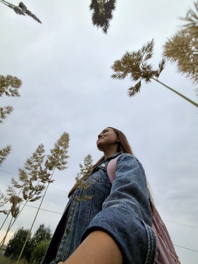 A young woman is captured from a low angle outdoors, looking up towards the sky. She is wearing a denim jacket and a pink backpack. Surrounding her are tall, dried grasses or reeds that reach upwards, framing her face and the cloudy sky. In the background, a line of trees and power lines suggest a semi-rural or suburban setting. The image appears to have been taken during the day under an overcast sky.