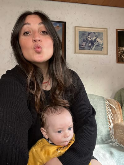 An adult female is depicted indoors, holding an infant. The female, with long dark hair, wearing a black ribbed sweater and a thin red necklace, faces the camera with pursed lips, indicating a "kiss" gesture. Her skin is light, and her eyes are dark. The infant, cradled against her, possesses light skin, blue eyes, and wispy dark hair, with a trace of saliva on its chin. The infant wears a yellow long-sleeved shirt bearing the partial word "MOMMY'S" and directs its gaze slightly to its left.

The background features a wall covered in white patterned wallpaper with subtle grey diagonal lines. Three framed items are mounted on this wall. From left to right: a partially visible frame containing what appears to be a Buddhist thangka; a central framed aerial photograph depicting multiple houses with red roofs and surrounding properties, characteristic of German residential architecture; and a partially visible frame on the right showing an aged or sepia-toned photograph.

The setting is a domestic interior, consistent with a living room in Wüstenrot, Germany, with the aerial photograph presumed to illustrate a view of Wüstenrot or its surrounding area. A light blue-green throw blanket with white fringe is draped over furniture on the right, where a section of a patterned upholstered chair or couch is also visible.